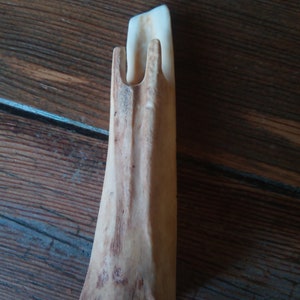 Puede incluir: Objeto de madera tallada de color marrón claro y forma única. El objeto tiene una base plana y una sección vertical con dos púas en la parte superior. La veta de la madera es visible.