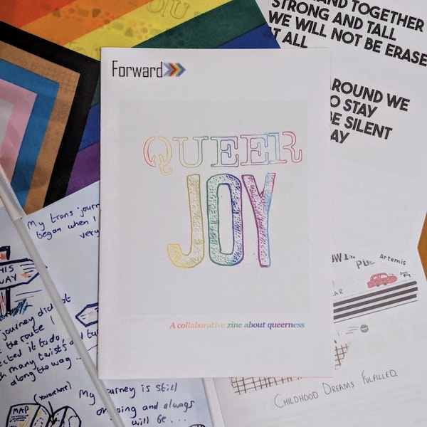 May include: A white zine titled "QUEER JOY" with rainbow-colored lettering. The text "A collaborative zine about queerness" is below the title. Other papers with handwritten text and colorful backgrounds surround the zine.