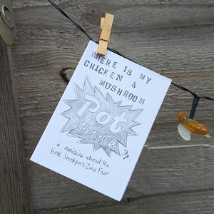 Puede incluir: Una minizine blanca, sujeta con una pinza de madera, presenta el título "WHERE IS MY CHICKEN & MUSHROOM Pot Novelle". La minizine trata sobre la First Stockport Zine Fair. También se ven una cadena de luces y un fondo de madera.