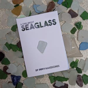 May include: A white zine titled "A Zine About Seaglass" with a piece of seaglass on the cover. The zine is surrounded by various pieces of sea glass in shades of green, blue, white, and brown. The text "BY PIPPYMAKESZINES" is at the bottom.
