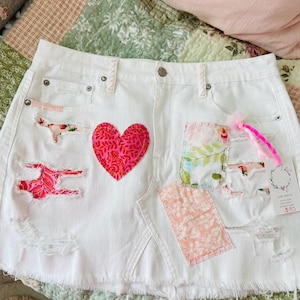 Peut inclure: Jupe en jean blanche avec détails déchirés et patchs en tissu colorés. Comprend un cœur rouge appliqué et des motifs floraux. La jupe a un ourlet effiloché et une fermeture boutonnée.