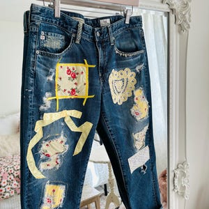 May include: Dark wash denim jeans with decorative patches. Patches include floral fabric squares, lace, and yellow ribbon accents. The jeans are cuffed at the ankles and hang from a pink hanger in front of a mirror.