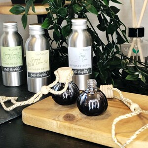 May include: Three silver metal diffuser refill bottles with labels that read "Blooper", "Blousey", and "Bijou". The bottles are on a wooden surface with two black glass car air fresheners hanging from a rope.