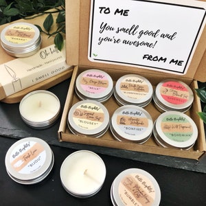 May include: A gift box containing six small round metal tins of soy wax candles with labels reading Belle Brightly and various scent names. The box lid features the text TO ME You smell good and you're awesome! FROM ME. Additional candles are placed outside.