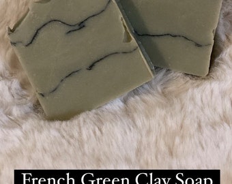 May include: Two bars of French Green Clay Soap with dark veining on a fluffy, light-colored surface. The soap has a light green hue and a textured, uneven edge. The text "French Green Clay Soap" is displayed in a black rectangle.