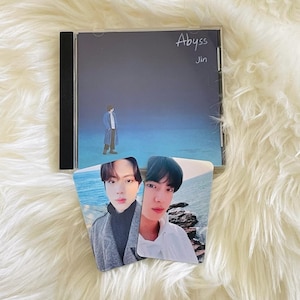 Puede incluir: Una caja de CD con un borde negro y un fondo azul y gris. El texto "Abyss" y "Jin" están escritos en la caja. Se muestran dos tarjetas de fotos con imágenes de dos hombres. Las tarjetas de fotos tienen un fondo azul y blanco.