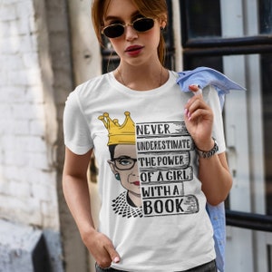 May include: White t-shirt with a graphic of Ruth Bader Ginsburg wearing a crown and holding books. The text on the shirt reads "Never underestimate the power of a girl with a book."