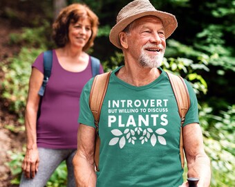 May include: A man wearing a green t-shirt with the text "Introvert but willing to discuss plants" and a white graphic of leaves. He is wearing a brown hat and is hiking in a forest.
