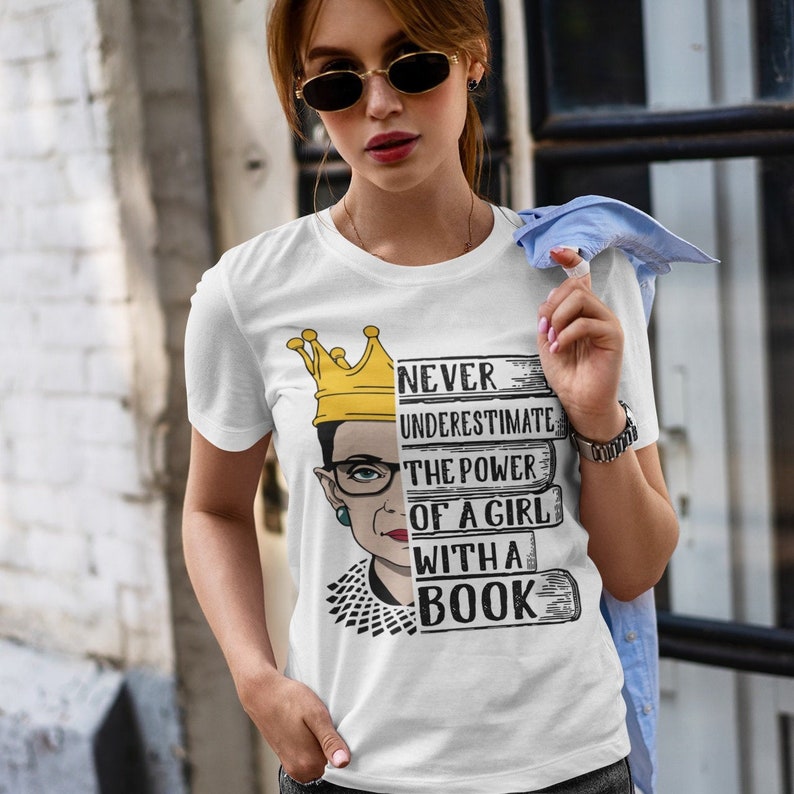 May include: White t-shirt with a graphic of Ruth Bader Ginsburg wearing a crown and holding books. The text on the shirt reads "Never underestimate the power of a girl with a book."