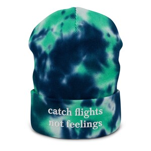 May include: A blue and green tie-dye beanie with the text "catch flights not feelings" embroidered in white.