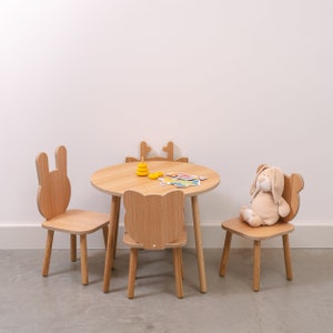 May include: A wooden children's table and chairs set with a light brown finish. The table has a round top and three legs. The chairs have bunny-shaped backs and four legs. A stuffed bunny sits on one of the chairs.