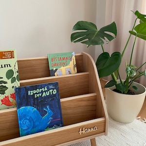 Puede incluir: Una estanter&iacute;a de madera con tres estantes, cada uno con un libro infantil diferente. La estanter&iacute;a est&aacute; etiquetada con "Hanna" y tiene un acabado de madera clara. Los libros se titulan "Laipni lugu muzeja", "Bistamais darzins" y "Izdoma sev auto".