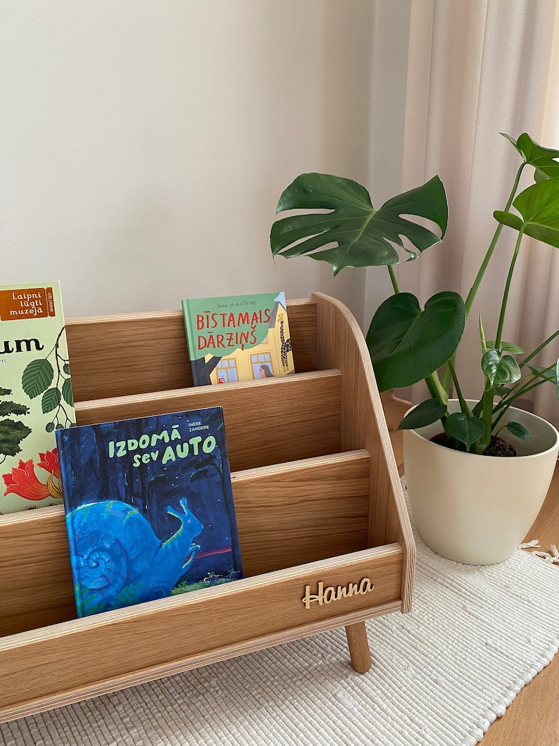 Puede incluir: Una estanter&iacute;a de madera con tres estantes, cada uno con un libro infantil diferente. La estanter&iacute;a est&aacute; etiquetada con "Hanna" y tiene un acabado de madera clara. Los libros se titulan "Laipni lugu muzeja", "Bistamais darzins" y "Izdoma sev auto".