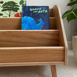 Puede incluir: Una estanter&iacute;a de madera con cuatro estantes, con dos libros para ni&ntilde;os. El libro de arriba se titula "Arboretum" y el libro de abajo se titula "Izdomā sev auto". La estanter&iacute;a tiene cuatro patas y est&aacute; hecha de madera marr&oacute;n claro.