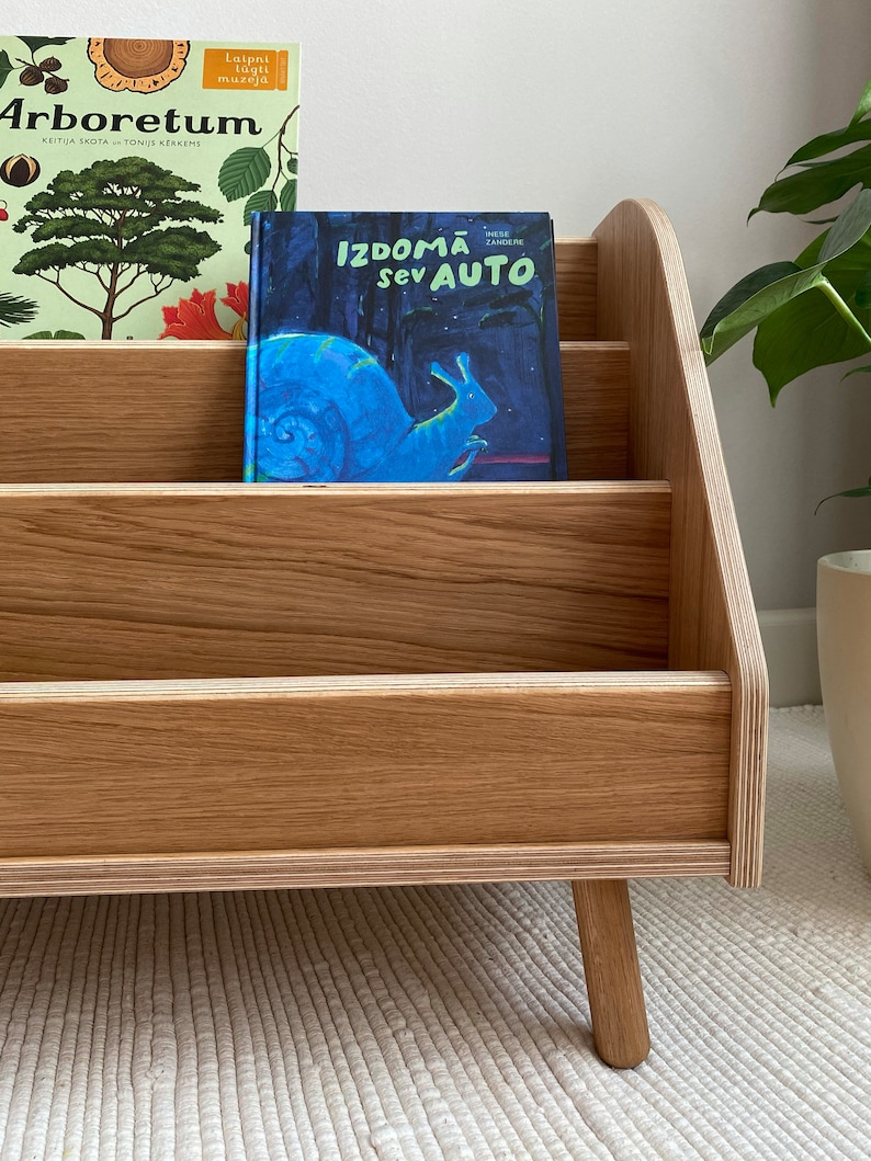 Puede incluir: Una estanter&iacute;a de madera con cuatro estantes, con dos libros para ni&ntilde;os. El libro de arriba se titula "Arboretum" y el libro de abajo se titula "Izdomā sev auto". La estanter&iacute;a tiene cuatro patas y est&aacute; hecha de madera marr&oacute;n claro.