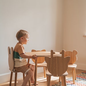 May include: A young child sits at a wooden table and chairs with animal-shaped backs. The table and chairs are made of light wood and have a natural finish. The child is wearing a brown and green striped shirt and brown shorts. The chairs are shaped like giraffes and have a light brown finish.