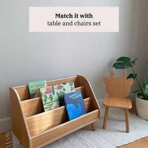 Puede incluir: Una estanter&iacute;a de madera con tres estantes, que contiene libros para ni&ntilde;os. La estanter&iacute;a tiene una parte superior curva y se apoya en cuatro patas. Una peque&ntilde;a silla de madera con un dise&ntilde;o de cabeza de ciervo se encuentra sobre una alfombra blanca delante de la estanter&iacute;a.