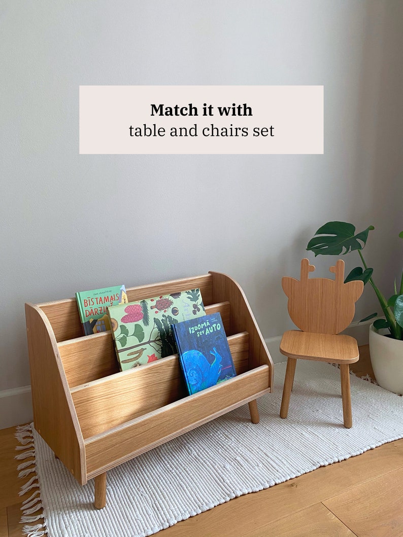 Puede incluir: Una estanter&iacute;a de madera con tres estantes, que contiene libros para ni&ntilde;os. La estanter&iacute;a tiene una parte superior curva y se apoya en cuatro patas. Una peque&ntilde;a silla de madera con un dise&ntilde;o de cabeza de ciervo se encuentra sobre una alfombra blanca delante de la estanter&iacute;a.