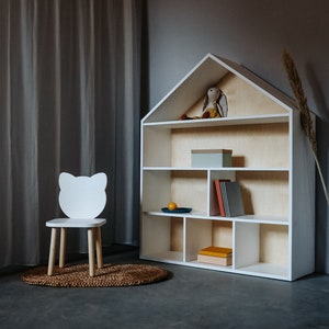 May include: White wooden bookshelf shaped like a house with shelves for books and toys. A white wooden chair with cat ears sits in front of the bookshelf.