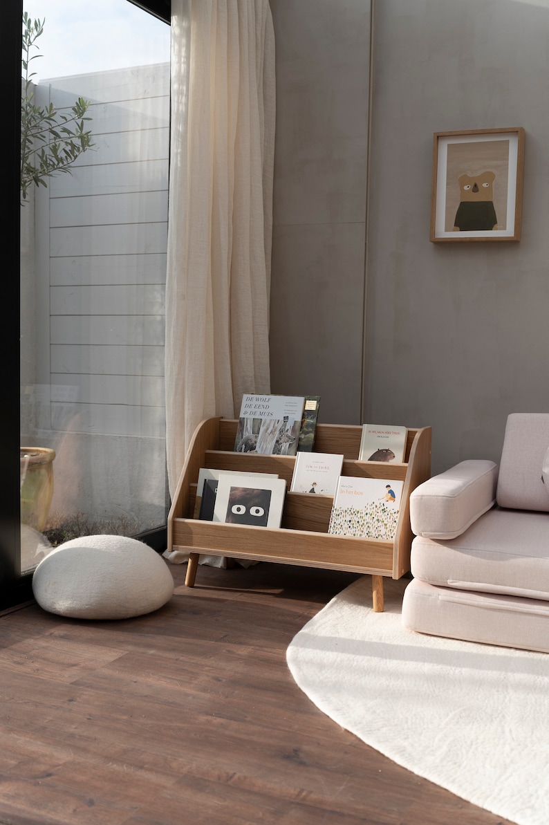 Puede incluir: Una estanter&iacute;a de madera con tres estantes, llena de libros. La estanter&iacute;a est&aacute; en una habitaci&oacute;n con una alfombra blanca, un puf blanco y un sof&aacute; rosa. La pared es de color gris claro.