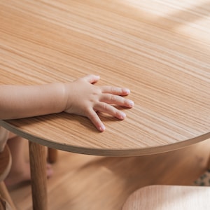 May include: A child's hand rests on a light wood table top. The table is round and has a light wood grain. The table is in focus, while the child's hand is slightly out of focus.