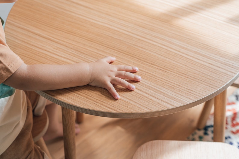 May include: A child's hand rests on a light wood table top. The table is round and has a light wood grain. The table is in focus, while the child's hand is slightly out of focus.