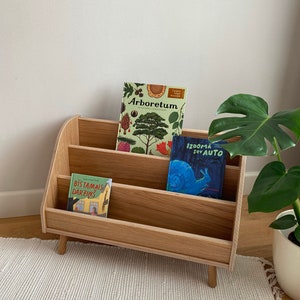 Puede incluir: Una estanter&iacute;a de madera con tres estantes, con tres libros para ni&ntilde;os. El estante superior tiene un libro titulado "Arboretum" con una cubierta verde e ilustraciones de &aacute;rboles. El estante del medio tiene un libro titulado "Izdoma sev auto" con una cubierta azul e ilustraciones de un caracol. El estante inferior tiene un libro titulado "Bistamals darzins" con una cubierta roja e ilustraciones de una casa. La estanter&iacute;a est&aacute; sobre una alfombra blanca.