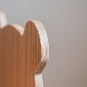 May include: Close-up of the back of a wooden chair with a rounded, ear-like shape. The chair is made of light brown plywood with visible layers.
