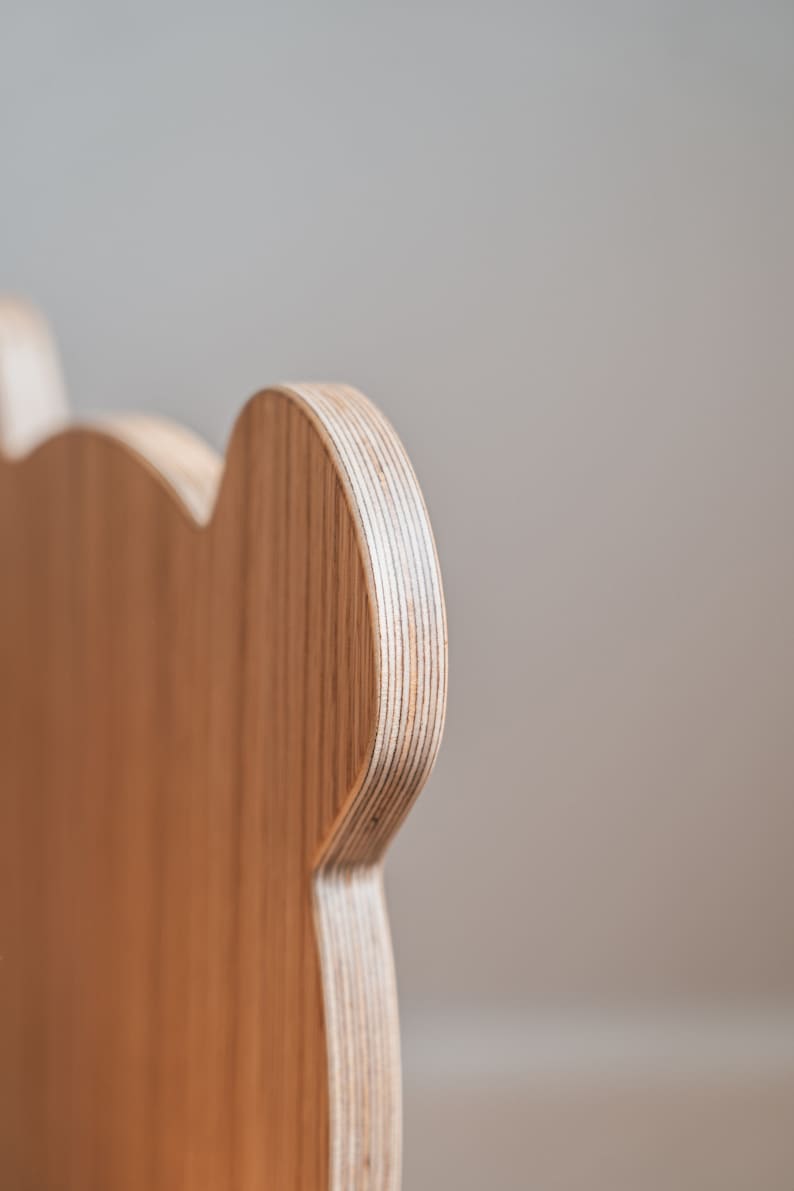 May include: Close-up of the back of a wooden chair with a rounded, ear-like shape. The chair is made of light brown plywood with visible layers.