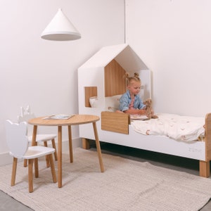 May include: A white wooden house-shaped bed with a white and brown mattress and a girl sitting on it. The bed has a small window and a wooden shelf. There is a white table and two white chairs with wooden legs in the foreground. The room has a white wall and a white ceiling.