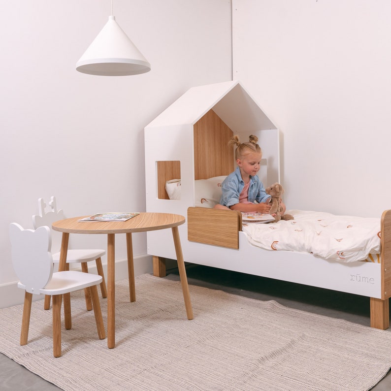 May include: A white wooden house-shaped bed with a white and brown mattress and a girl sitting on it. The bed has a small window and a wooden shelf. There is a white table and two white chairs with wooden legs in the foreground. The room has a white wall and a white ceiling.