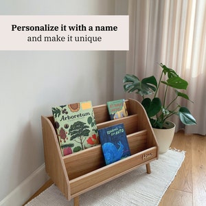 Puede incluir: Una estanter&iacute;a de madera con tres estantes, con una placa de nombre personalizada que dice "Hanna". La estanter&iacute;a est&aacute; llena de libros para ni&ntilde;os, incluyendo "Arboretum", "Bistamais Darzins" y "Izdoma Sev Auto".