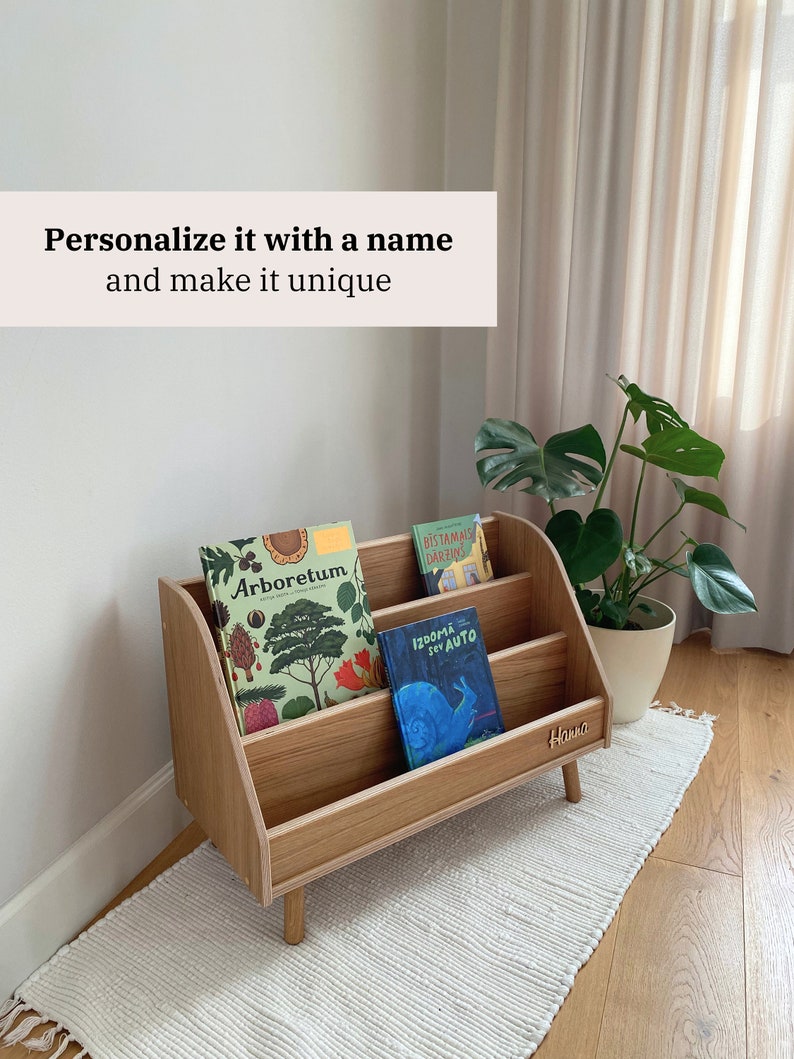 Puede incluir: Una estanter&iacute;a de madera con tres estantes, con una placa de nombre personalizada que dice "Hanna". La estanter&iacute;a est&aacute; llena de libros para ni&ntilde;os, incluyendo "Arboretum", "Bistamais Darzins" y "Izdoma Sev Auto".