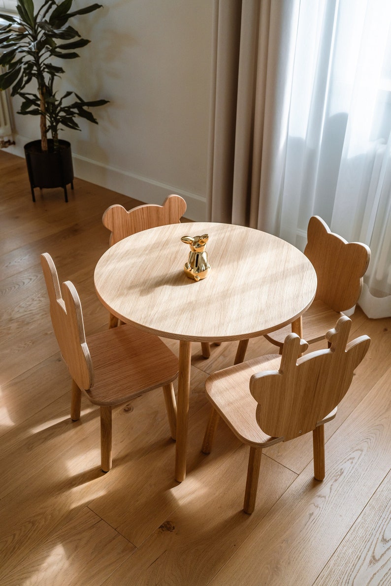 May include: A wooden children's table and four chairs with animal-shaped backs. The table is round and has a light wood finish. The chairs are also light wood and have a variety of animal shapes, including a bear, a deer, and a rabbit. A gold figurine sits in the center of the table.