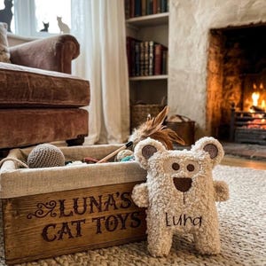 May include: A wooden storage crate with the text LUNA'S CAT TOYS printed on the front, containing various items. Beside the crate sits a cream-coloured, textured plush bear toy with the name Luna embroidered in brown thread on its chest.