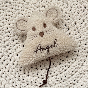 May include: A triangular, cream-coloured, sherpa-textured mouse ornament featuring embroidered brown eyes, a nose, whiskers, and the name Angel stitched in brown script. A thin brown cord extends from the bottom of the plush mouse figure.