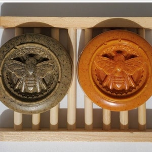 Puede incluir: Dos jabones redondos con un diseño de abeja en un jabonera de madera. El jabón de la izquierda es verde y el jabón de la derecha es naranja.
