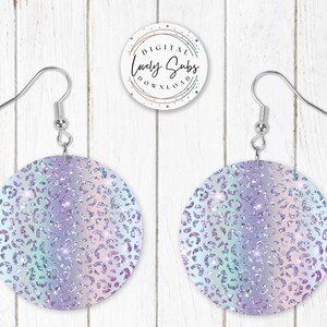 May include: A pair of silver hoop earrings with a round, iridescent blue and purple glitter design featuring a leopard print pattern. The earrings are on a white wooden background.
