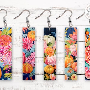May include: Five pairs of rectangular earrings with floral and pumpkin designs. Each earring features a different colorful arrangement of flowers and pumpkins against a dark blue background. The earrings have silver-colored hooks.