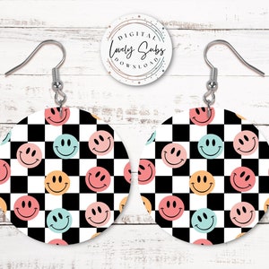 May include: A pair of circular earrings with a black and white chequered design. Each earring displays colourful smiley faces in pink, blue, and yellow. The earrings have silver hooks and a white background.