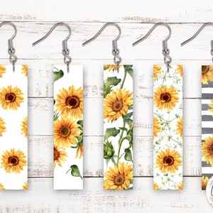May include: Five pairs of dangle earrings with different sunflower designs. The earrings are silver and have a rectangular shape. The designs include a white background with sunflowers, a white background with sunflowers and green leaves, a white background with small white flowers and sunflowers, and a black and white striped background with sunflowers.