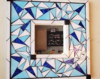 Könnte beinhalten: Ein quadratischer Spiegel mit einem Mosaikrahmen aus blauen, weißen und schwarzen Glasfliesen. Der Rahmen besteht aus kleinen, geometrischen Formen. Der Spiegel spiegelt einen Raum mit einem Bücherregal und anderen Gegenständen wider.