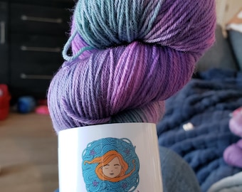 May include: A skein of hand-dyed yarn in shades of purple, blue, and teal. The yarn is wrapped around a white label with the text "Yarn for the Soul" and the website address "www.yarnforthesoul.co.uk".