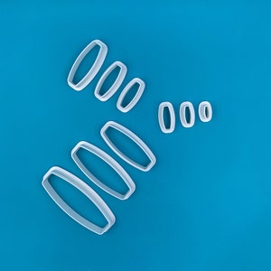 Puede incluir: Un conjunto de ocho cortadores de galletas de plástico blanco con forma de óvalos de diferentes tamaños. Los cortadores están dispuestos en diagonal sobre un fondo azul.