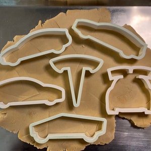 May include: Five white plastic cookie cutters shaped like Tesla cars and the Tesla logo on a brown surface.