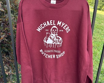 May include: A maroon sweatshirt with a white graphic featuring the text "MICHAEL MYERS BUTCHER SHOP" and an illustration of Michael Myers. The text includes "EST 1978" and "ALWAYS FRESH". The sweatshirt is hanging on a black hanger.