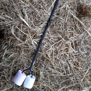 Könnte beinhalten: Ein schwarzer Metall-Röststab mit Schlaufengriff und zwei Zinken, die ein rosa und ein weißes Marshmallow halten. Der Stab liegt auf einem Bett aus Heu.
