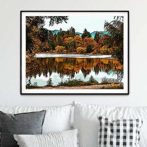 May include: A framed print depicting a lake reflecting a forest with autumn foliage. The trees are a mix of green, yellow, orange, and red. The water is calm and still, mirroring the colours of the trees.