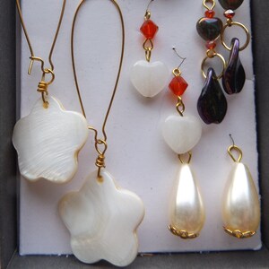 May include: A collection of earrings displayed in a gray box. The earrings feature various designs, including gold-toned geometric shapes, red and white heart-shaped beads, and pearly white teardrops. Some earrings have gold-colored wire accents and white flower-shaped pendants.