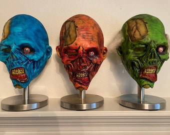 May include: Three Halloween masks on display stands. The masks are made of painted plaster and feature realistic zombie faces. The masks are blue, red, and green.
